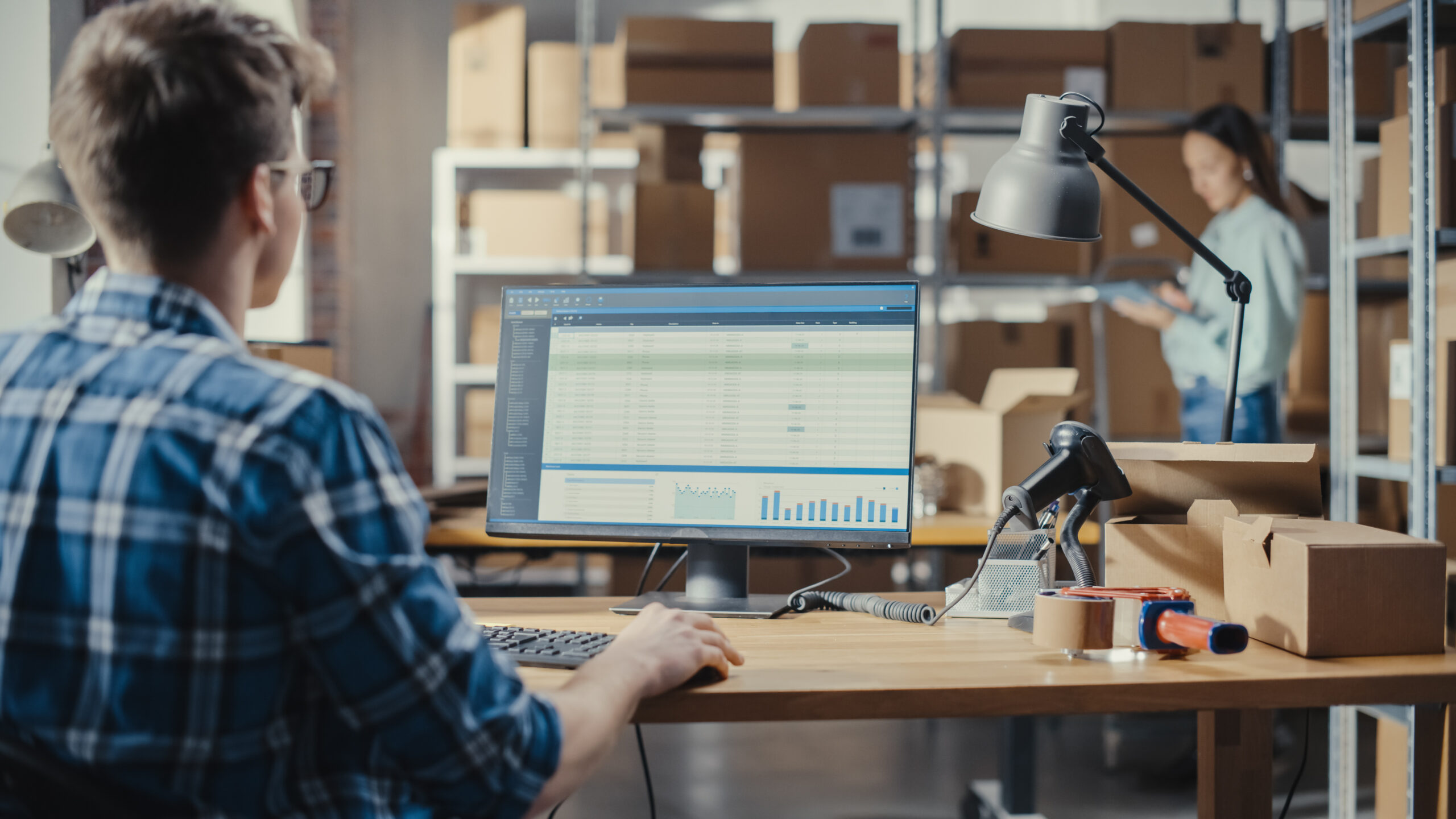 Warehouse office worker checking inventory records on a desktop screen beside packing supplies and stock shelves