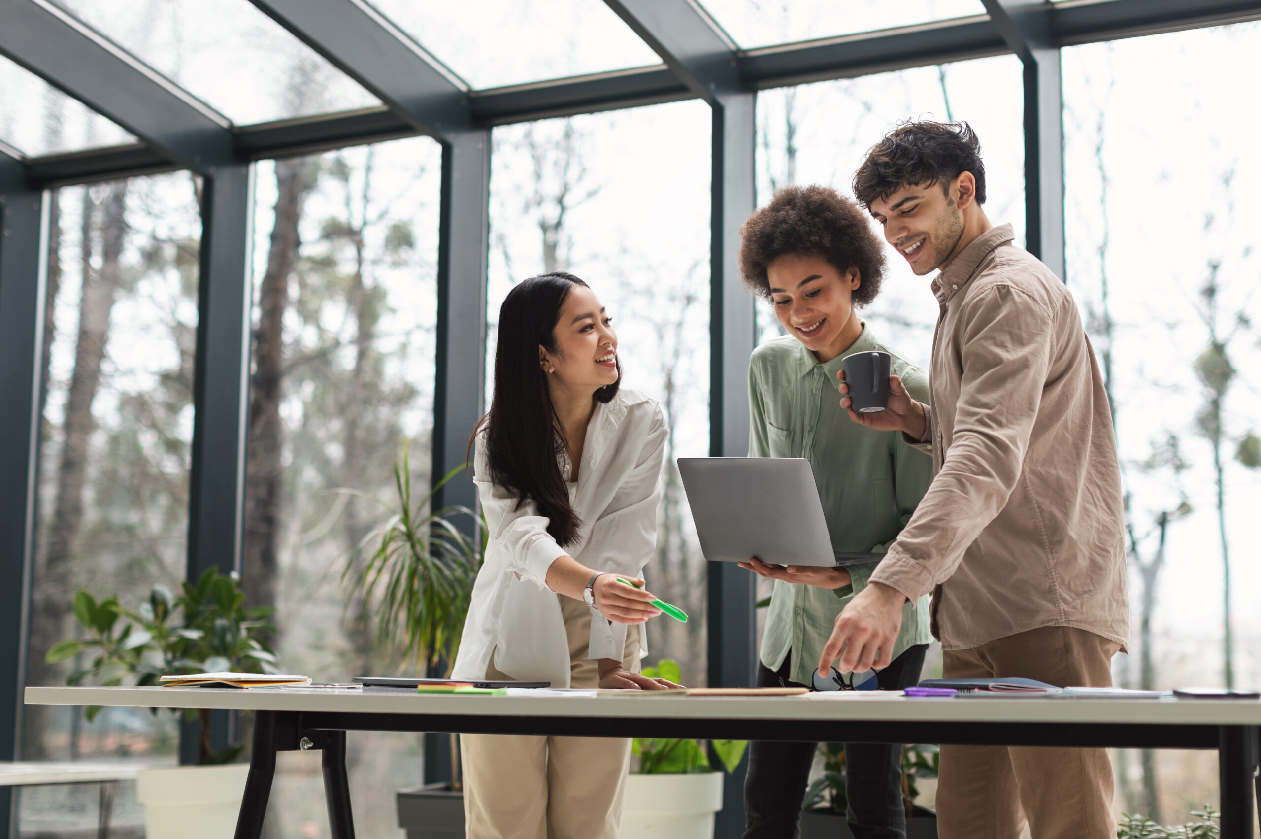 Three colleagues reviewing a laptop together in a bright glass-roofed office