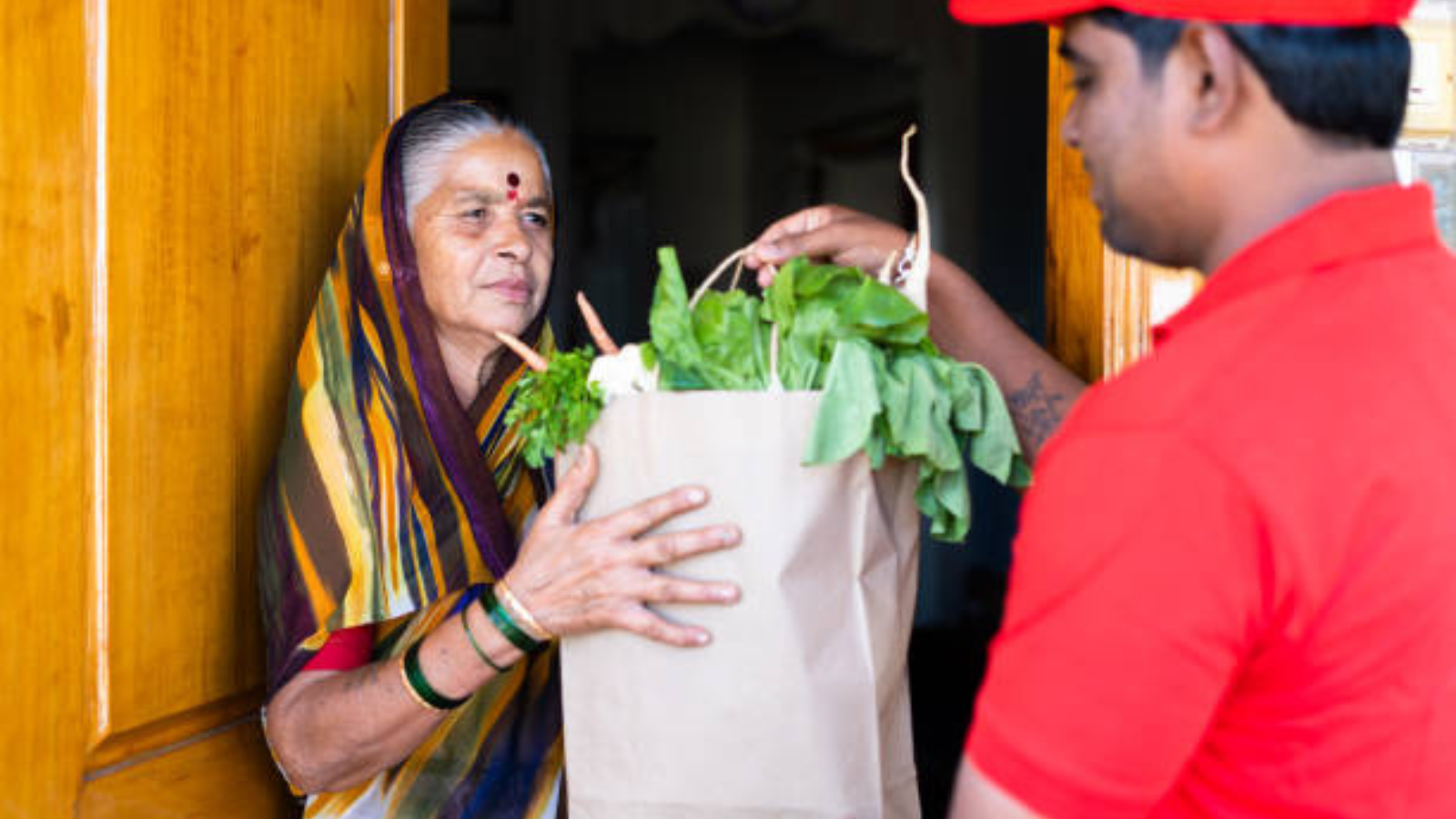 Quick commerce grocery delivery in urban India, showing a last-mile delivery executive handing fresh vegetables to a customer at her doorstep.