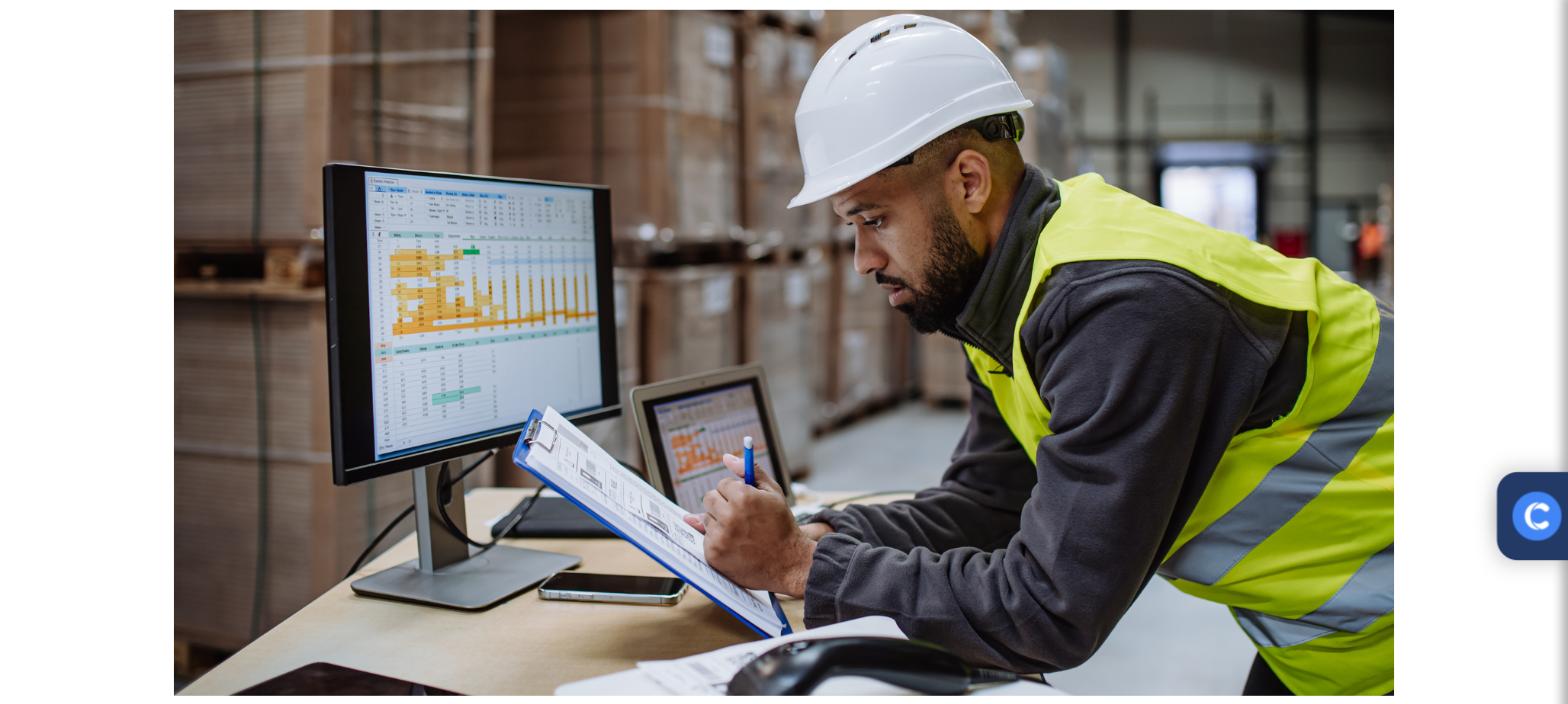 Warehouse worker in a hard hat reviewing multi-warehouse stock levels on a monitor and tablet