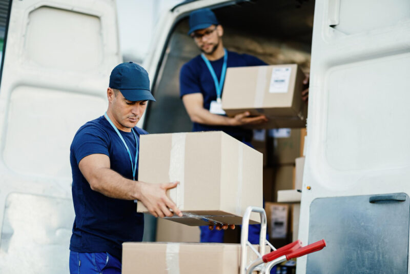 Un joven mensajero y su colega descargando cajas de cartón de una furgoneta de reparto.